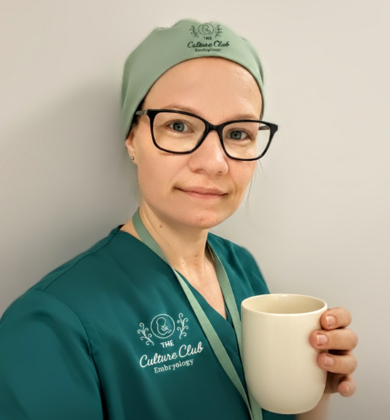 Dr Katie Finch in The Culture Club Embryology scrubs and theatre cap, holding a mug.